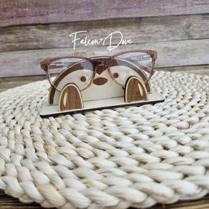 May include: A light-colored wooden glasses stand featuring a stylized animal face design with two dark brown wooden paws at the base. The stand holds a pair of clear-lens glasses with thin, light brown frames. The text Falcon + Dove is visible.