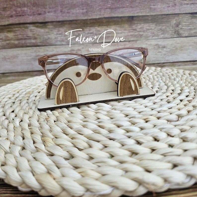 May include: A light-colored wooden glasses stand featuring a stylized animal face design with two dark brown wooden paws at the base. The stand holds a pair of clear-lens glasses with thin, light brown frames. The text Falcon + Dove is visible.