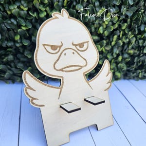 May include: A light-colored wooden phone stand shaped like a cartoon duck with an angry expression. The duck has etched outlines, two small protruding supports for a device, and the text Falcon + Dove STUDIO is visible in the upper right corner.
