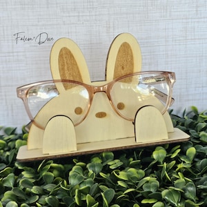 May include: A light-colored wooden glasses stand shaped like a rabbit with two tall ears, two small circular eyes, and a small oval nose. The stand features two front-facing tabs that hold a pair of light brown-rimmed glasses in place.