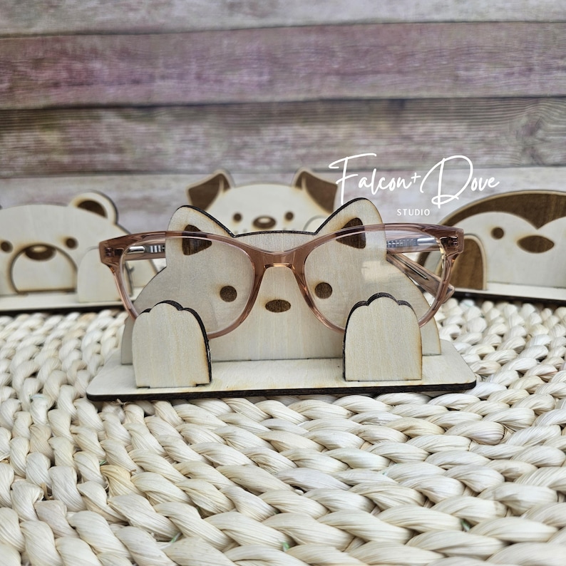 May include: A light-colored wooden glasses holder shaped like a stylized animal with two raised paws. The holder features engraved facial details and is currently displaying a pair of translucent pink-tinted glasses. The text Falcon + Dove Studio is visible.