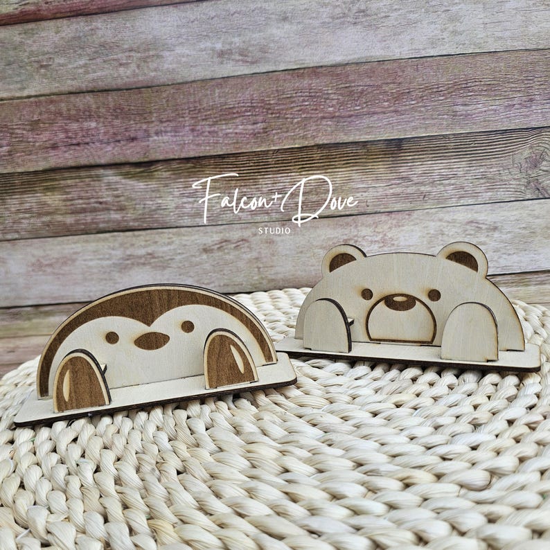 May include: Two wooden phone stands featuring animal designs. One stand is shaped like a brown and light wood penguin, and the other is a light wood bear. Both stands have a flat base and a cutout design to hold a mobile device securely.