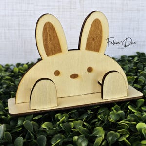 May include: A light-colored wooden stand shaped like a rabbit with two tall ears, two small circular eyes, and an oval nose. The stand features two rounded front-facing paws and the text Falcon + Dove Studio printed on the right side.