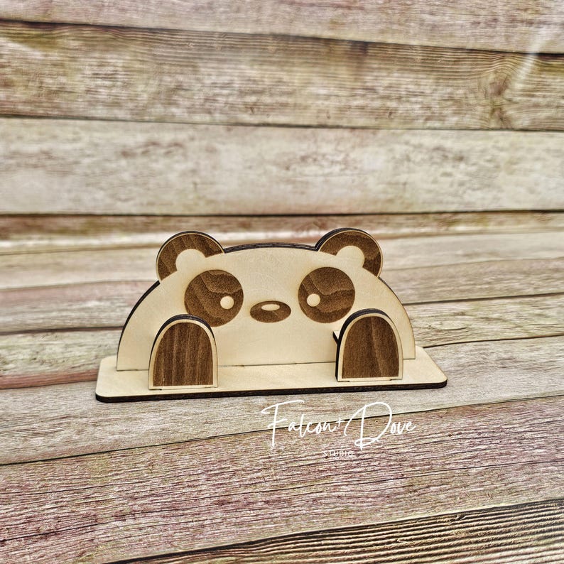 May include: A light-colored wooden phone stand shaped like a panda bear with dark brown accents on the ears, eyes, and paws. The stand features a horizontal base and a vertical back piece with a cutout design. The text Falcon and Dove Studio is visible.