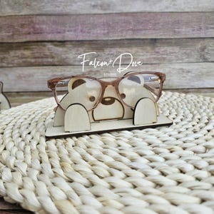 May include: A light-colored wooden eyeglass stand shaped like a bear with two rounded ears and a snout, holding a pair of translucent brown-rimmed glasses. The text Falcon + Dove STUDIO is printed in white above the bear-shaped wooden stand.