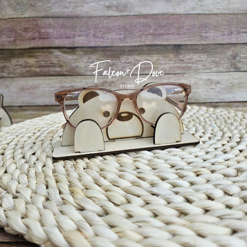 May include: A light-colored wooden eyeglass stand shaped like a bear with two rounded ears and a snout, holding a pair of translucent brown-rimmed glasses. The text Falcon + Dove STUDIO is printed in white above the bear-shaped wooden stand.