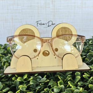May include: A light-colored wooden glasses stand shaped like a bear face with rounded ears, a small nose, and whiskers. The stand features the text Falcon + Dove Studio and holds a pair of translucent peach-colored eyeglasses on its front.