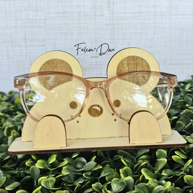 May include: A light-colored wooden glasses stand shaped like a bear face with rounded ears, a small nose, and whiskers. The stand features the text Falcon + Dove Studio and holds a pair of translucent peach-colored eyeglasses on its front.