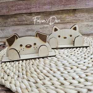 May include: Two light-colored wooden phone stands featuring laser-cut designs of a dog and a cat. The dog stand has dark brown ears and a snout, while the cat stand has pointed ears. Both figures have paws resting on a flat base. Text reads Falcon+Dove STUDIO.