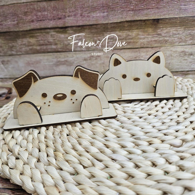 May include: Two light-colored wooden phone stands featuring laser-cut designs of a dog and a cat. The dog stand has dark brown ears and a snout, while the cat stand has pointed ears. Both figures have paws resting on a flat base. Text reads Falcon+Dove STUDIO.