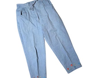 May include: Light blue denim trousers featuring an elastic waistband with a drawstring, two side pockets, and colourful embroidered floral accents with small red and blue circular details along the bottom hem of each trouser leg.