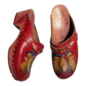Puede incluir: Par de zuecos de madera rojos con diseños florales pintados. Los zuecos tienen un tacón alto y una correa de cuero. El diseño floral incluye acentos amarillos, verdes y azules. Los zuecos son de estilo tradicional.
