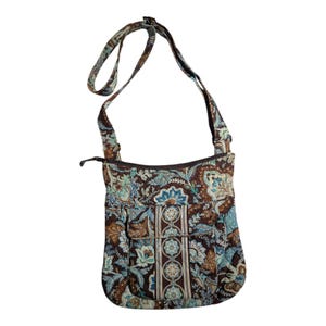 May include: A brown crossbody bag with a floral pattern in shades of blue, teal, and tan. The bag has a long adjustable strap, a zippered main compartment, and two front pockets. The bag is made of a quilted fabric.