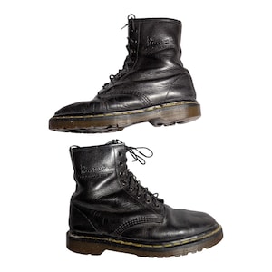 Puede incluir: Dos botas negras de cuero con cordones. Las botas muestran signos de desgaste con pliegues y rozaduras. Las suelas son de color marrón. Las botas son de estilo clásico, adecuadas para varios conjuntos.