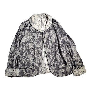 May include: A long-sleeve jacket featuring a dark gray and white floral paisley pattern. The garment has a shawl collar, a front opening, and folded cuffs. The interior lining and collar underside are white with a dark gray floral print.