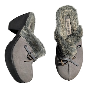 Pode incluir: Tamancos cinzentos de camurça sintética com acabamento em pele sintética. Os tamancos têm um detalhe de laço na frente e um salto cunha preto. A marca "Skechers" é visível no interior do sapato.