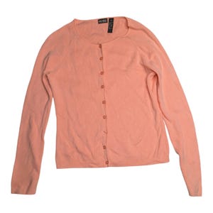 Pode incluir: Um cardigan de cor pêssego claro com mangas compridas e decote redondo. O suéter tem uma frente abotoada com sete botões correspondentes. A etiqueta na parte de trás do pescoço diz "CRO-SHEE".