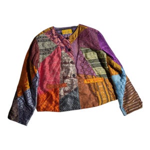 May include: A long-sleeve jacket featuring a multi-coloured patchwork design with various textured fabrics in shades of purple, orange, pink, blue, and gold. The jacket has a round neckline, a front button closure, and two horizontal pockets.