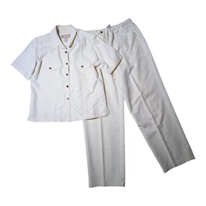 May include: A two-piece cream-colored outfit. The short-sleeved button-down shirt has two chest pockets and brown buttons. The matching pants are straight-legged. The outfit is made of a lightweight fabric.