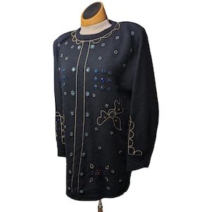 Può includere: Maglione nero con finiture e abbellimenti dorati. Il maglione presenta accenti blu e trasparenti simili a gioielli e disegni di fiocchi dorati. Lo scollo e i bordi sono rifiniti con filo d'oro. Il maglione è esposto su un manichino.