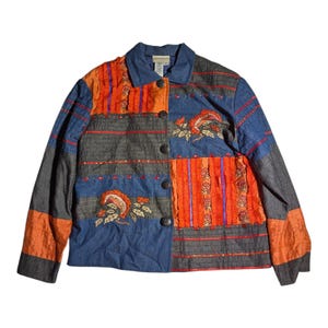 May include: A long-sleeve button-down jacket featuring a patchwork design of blue denim, charcoal gray fabric, and bright orange textured panels. The garment includes decorative red stitching, ruffled orange trim, and embroidered floral motifs.