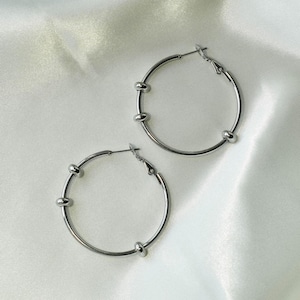 May include: A pair of silver hoop earrings with small round accents. The earrings are on a white satin background.
