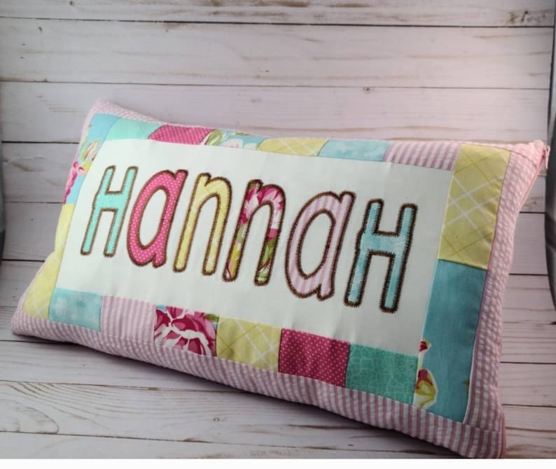 May include: A pink and white patchwork pillow with the name "Hannah" embroidered in colorful letters.