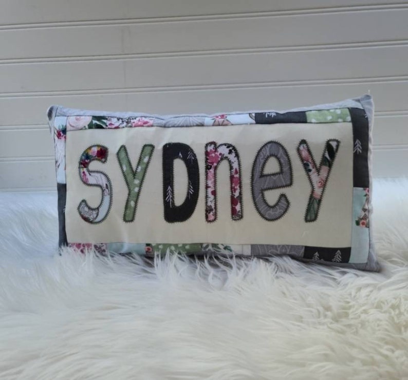 May include: A patchwork pillow with the name "sydney" spelled out in large, colorful letters. The pillow is made of various fabrics, including floral prints, geometric patterns, and solid colors.