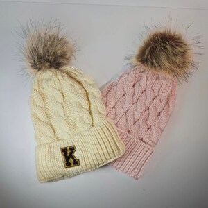 May include: Two knitted winter hats with faux fur pom poms. One hat is white with a black and gold "K" embroidered on the front. The other hat is pink.