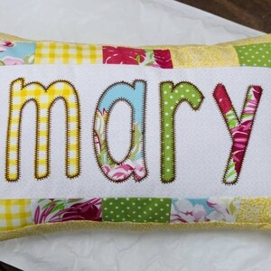 May include: A yellow and white patchwork pillow with the name "mary" embroidered in brown on the front.