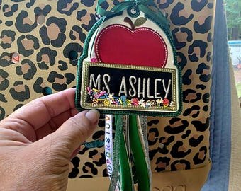May include: A personalized teacher bag tag with a red apple design and the name "Ms. Ashley" on a black background. The tag is adorned with colorful embellishments and green ribbons, hanging from a leopard print tote bag.