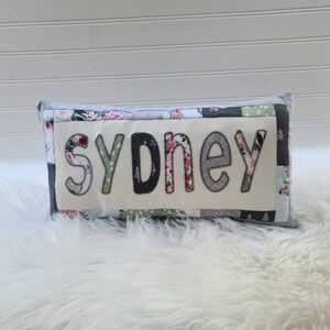 May include: A white pillow with a patchwork design featuring floral and geometric patterns. The word "Sydney" is spelled out in large, colorful letters on the front of the pillow.