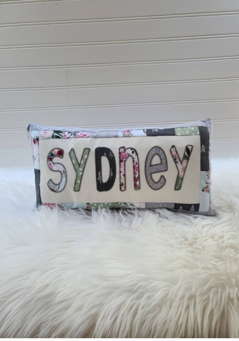 May include: A white pillow with a patchwork design featuring floral and geometric patterns. The word "Sydney" is spelled out in large, colorful letters on the front of the pillow.