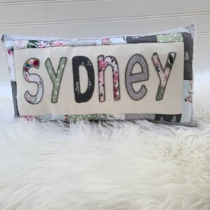 May include: A white pillow with a patchwork design featuring floral and geometric patterns. The word "sydney" is spelled out in large, gray letters on the front of the pillow.