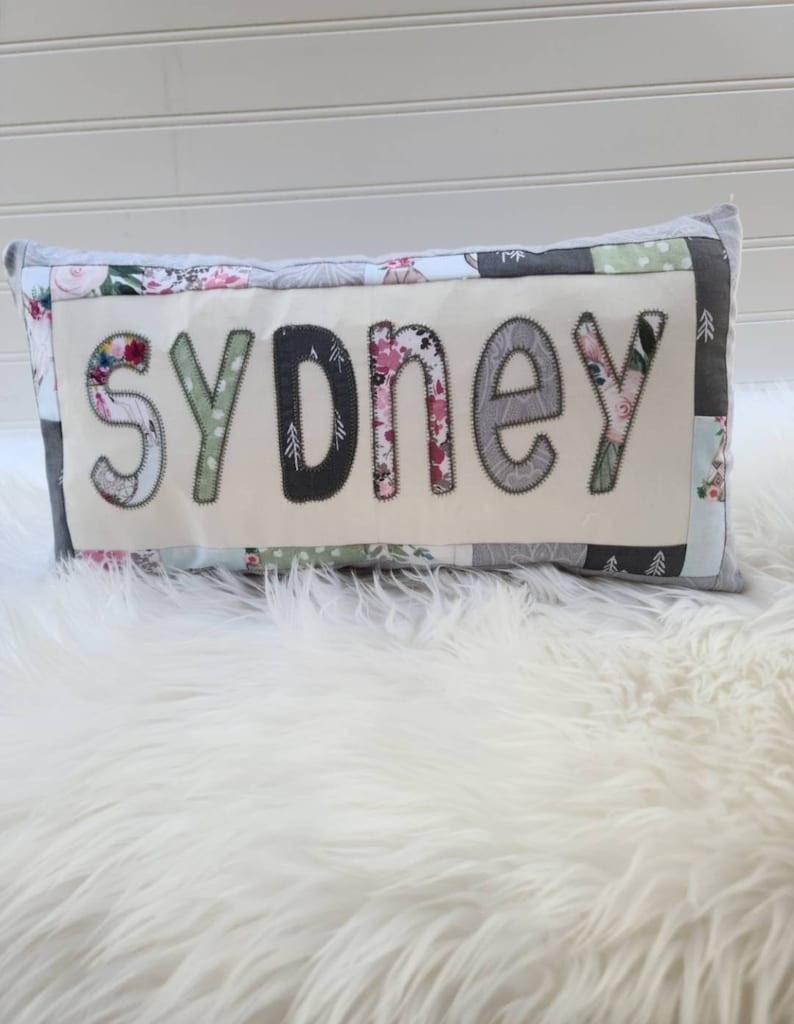 May include: A white pillow with a patchwork design featuring floral and geometric patterns. The word "sydney" is spelled out in large, gray letters on the front of the pillow.