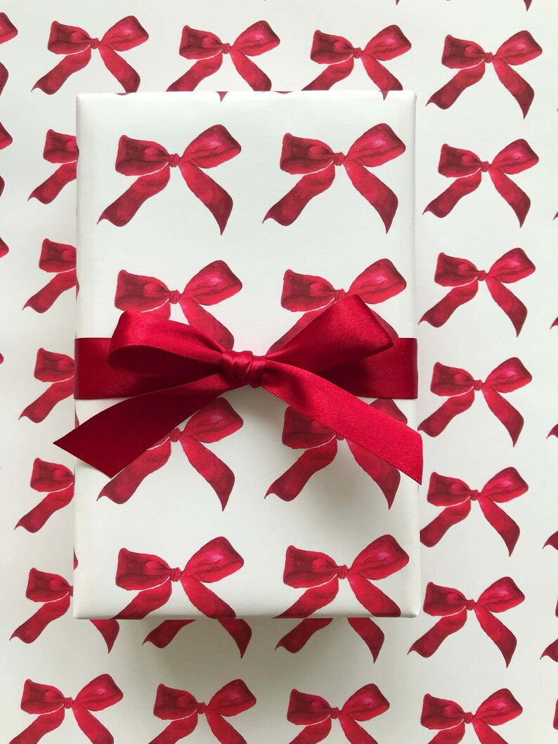 May include: White gift wrap with a repeating pattern of red watercolor bows. A red satin ribbon is tied around the gift wrap.