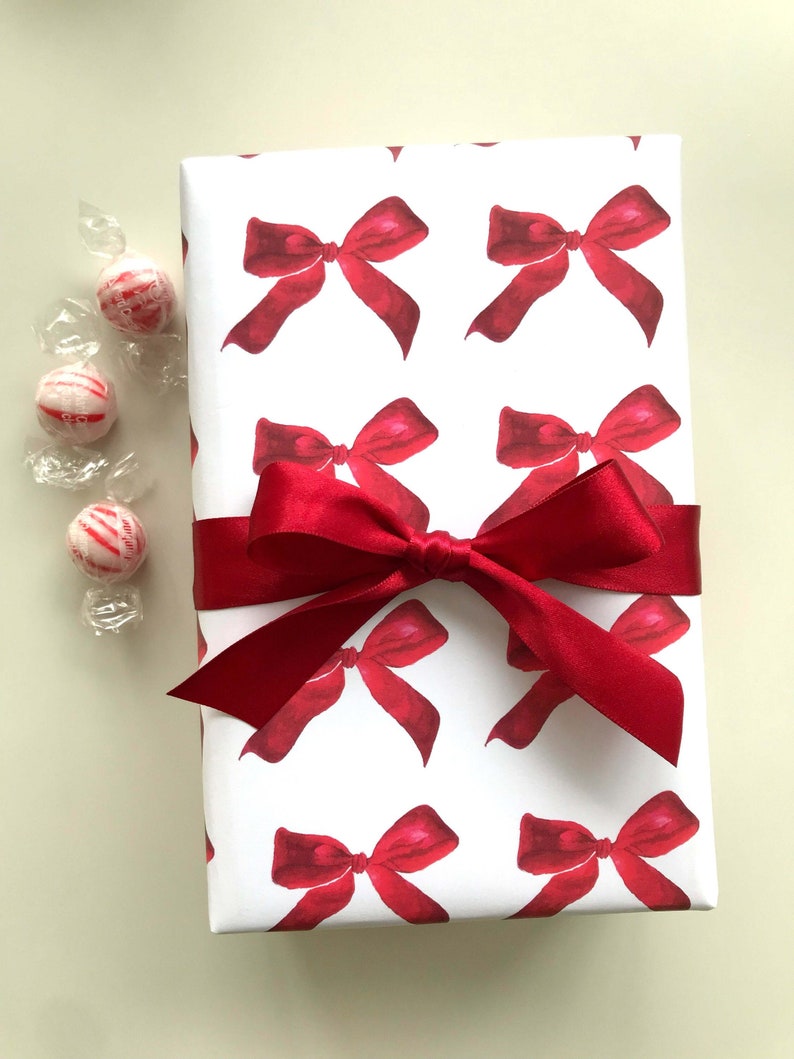 May include: White gift wrap with a repeating pattern of red watercolor bows. A red satin ribbon is tied around the gift wrap.