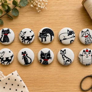 May include: Ten circular fabric buttons featuring black and white illustrations of cats in various poses. Each button displays a unique feline character, some with small red accents on their collars or heads, arranged in two rows on a wooden surface.