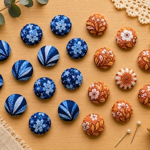May include: A collection of round fabric-covered buttons arranged on a surface. The buttons feature various patterns, including blue and white floral and leaf designs, as well as orange and white botanical motifs with intricate stitching details.