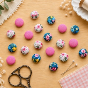 May include: A collection of round fabric-covered buttons in solid pink, white with pink floral patterns, and blue with multi-coloured floral prints. The buttons are arranged on a flat surface alongside metal sewing scissors and two straight pins.