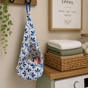 May include: A fabric bag with a blue and white floral pattern, suspended by a blue carabiner clip. The bag is filled with items. A sign reading "LAUNDRY room" is visible, alongside folded towels and a washing machine in a laundry room setting.