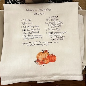 May include: A white tea towel with a handwritten pumpkin bread recipe. The recipe includes ingredients like flour, sugar, butter, eggs, and pumpkin pie filling. The recipe also includes instructions to bake at 325 degrees Fahrenheit for one hour in a greased baking dish. The tea towel is decorated with a watercolor illustration of pumpkins and flowers.