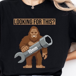 May include: Black t-shirt featuring a cartoon Bigfoot holding a 10mm wrench. The text "LOOKING FOR THIS?" is above the Bigfoot graphic. The shirt is worn by a person in blue jeans.
