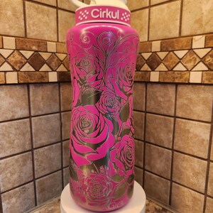 Pu&ograve; includere: Una borraccia rosa isolata con coperchio e manico bianchi. La bottiglia &egrave; decorata con un motivo a rose e la scritta "Cirkul". La bottiglia &egrave; su un supporto bianco.