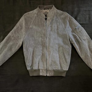 May include: A grey suede bomber jacket featuring a full-length front zip, a stand-up collar, and ribbed knit cuffs and hem. The jacket includes two side pockets and a small zipped utility pocket on the upper left sleeve with long sleeves.