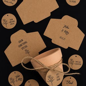 May include: A terracotta plant pot wrapped in twine with a round brown paper tag reading LET LOVE GROW, surrounded by various brown paper tags featuring floral illustrations and text including FROM OUR SHOWER TO YOURS, JORDAN & RILEY, and THANK YOU.