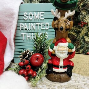May include: A Christmas figurine of Santa Claus riding on top of two reindeer. The reindeer are brown and have red noses. Santa is wearing a red suit with white trim and a white beard. The figurine is on a wooden base. The background is a green and red Christmas pattern.