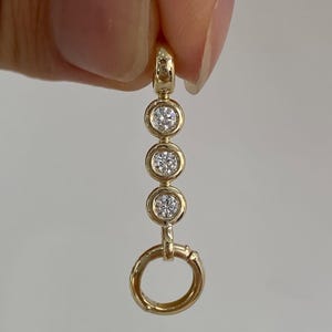 Puede incluir: Un colgante de oro con una fila vertical de tres piedras redondas, claras y facetadas, engastadas en biseles de oro pulido, rematado con una pequeña anilla de oro y terminado con un aro de oro en la parte inferior, de diseño lineal.