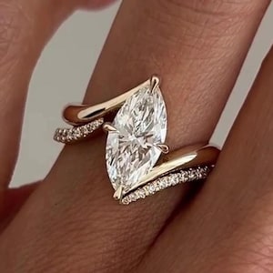 May include: A gold engagement ring featuring a marquise-cut diamond. The band has a split shank design, with small diamonds set along the band. The ring is displayed on a finger.