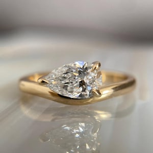 Puede incluir: Un anillo de oro con un diamante en forma de pera. El diamante es transparente y facetado, reflejando la luz. La banda del anillo es de oro pulido. El anillo está sobre una superficie reflectante.